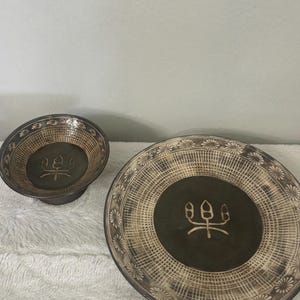 May include: A decorative ceramic bowl and plate set. The bowl and plate are dark green with a beige woven pattern and floral border. Each piece has a central design of a stylized symbol.
