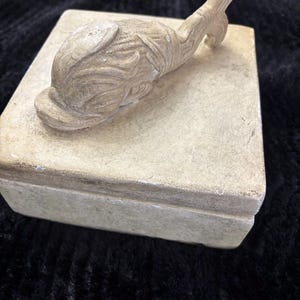 May include: A beige, rectangular box with a sculpted, stylized bird figure on top. The bird is light beige with carved details. The box and bird appear to be made of a stone-like material. The background is a dark, textured fabric.