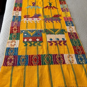 May include: A vibrant yellow textile with intricate embroidered designs. The rectangular piece features repeating patterns of birds and animal figures in red, green, blue, and white. The edges are adorned with a fringe.