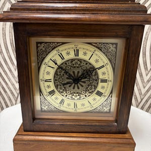 May include: A square wooden mantel clock with a dark brown finish. The clock face features a cream-colored background with black Roman numerals, a central floral pattern, and ornate corner decorations. It has black hands and a metal handle on top.