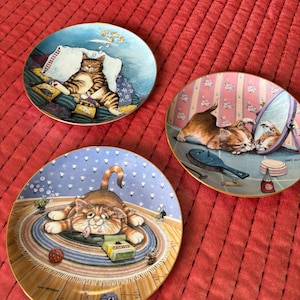May include: Three decorative plates featuring cat illustrations. One plate shows a cat sleeping on pillows, another shows a cat looking in a mirror, and the third shows a cat with a catnip box. Each plate has a gold rim.