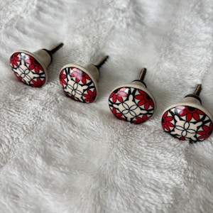 May include: Four decorative cabinet knobs with a floral design. Each knob features a cream-colored base with a pattern of red flowers and black accents. The knobs are arranged on a white, textured surface.