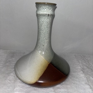 May include: A handcrafted ceramic vase with a unique color-blocked design. The vase features a slender, textured neck in shades of gray and a bulbous base divided into sections of white, tan, brown, and dark gray. The vase is approximately 10 inches tall.