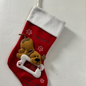 May include: Red Christmas stocking with a white cuff and a felt dog design. The dog is holding a bone. The text "Dear Santa" and "I've been a very good doggy all year" are embroidered on the stocking, along with snowflakes.