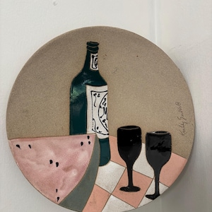 May include: A round ceramic plate featuring a hand-painted scene of a dark green wine bottle with a white label, two black wine glasses, and a slice of pink watermelon with black seeds, all set against a checkered pink and white surface.