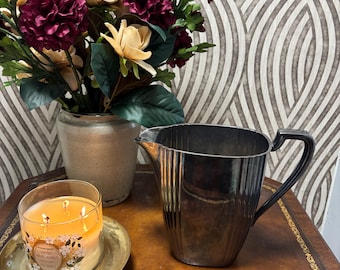 May include: A silver pitcher with a black handle sits on a brown surface next to a lit candle in a glass jar on a gold-colored plate. A vase of flowers with burgundy and cream-colored blooms is in the background.