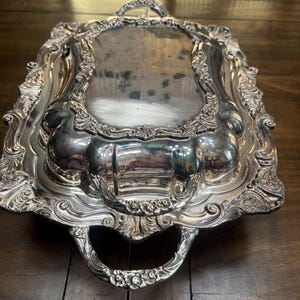 May include: A silver-colored, rectangular serving tray with ornate detailing. The tray features a reflective surface and decorative handles on each end. The edges are embellished with intricate scrollwork and floral patterns, creating an elegant aesthetic.
