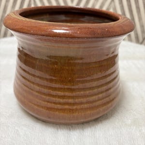 May include: A small, round ceramic pot with a glossy, warm brown glaze. The vessel features a wide, flared rim and a series of horizontal, subtle ridges encircling the body, creating a textured appearance across the smooth, reflective surface.