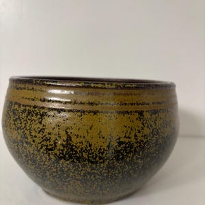 May include: A handcrafted ceramic bowl with a speckled, textured surface. The bowl features a dark brown rim and a gradient of olive green to black, creating a rustic aesthetic. This pottery piece is ideal for serving or display.