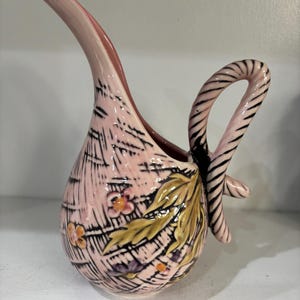 May include: A pink ceramic pitcher with a long, curved spout and a striped handle. The pitcher features a textured pattern and is decorated with floral and leaf motifs in yellow, orange, and purple.