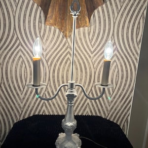 May include: A white candelabra lamp featuring two light sockets with flame-shaped bulbs, a central vertical rod with a decorative finial, and an ornate, scrolled base. The lamp has two curved arms extending from the central column.
