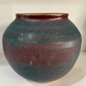 May include: A handcrafted ceramic vase with a rounded body and a slightly flared rim. The vase features a color gradient, transitioning from a deep burgundy at the base to a teal-green midsection, and a dark red rim. The textured surface adds to its rustic charm.