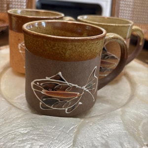 May include: Three ceramic mugs with a matte brown finish and a speckled tan rim. Each mug features a white line-art illustration of a fish on the side. The mugs have a sturdy handle and a cylindrical shape, showcasing a consistent earthy color palette.