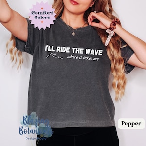 May include: A dark gray t-shirt with the text "I'LL RIDE THE WAVE where it takes me" and a wave graphic in white.