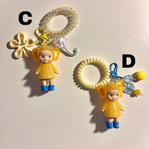 May include: Two keychains featuring yellow figures in elephant costumes, blue boots, and attached to spiral hair ties. One keychain has a flower charm, the other has a cluster of beads.