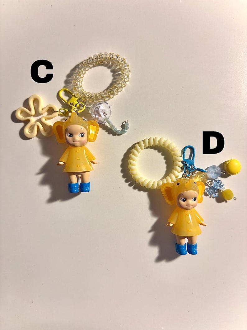 May include: Two keychains featuring yellow figures in elephant costumes, blue boots, and attached to spiral hair ties. One keychain has a flower charm, the other has a cluster of beads.