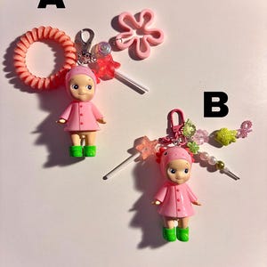 May include: Two keychains featuring small doll figures in pink coats and green boots. One keychain has a pink hair tie and a pink flower charm. The other has a beaded charm with a star and fish. Both have a lollipop charm.
