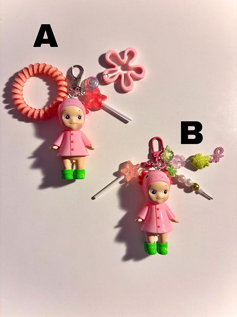 May include: Two keychains featuring small doll figures in pink coats and green boots. One keychain has a pink hair tie and a pink flower charm. The other has a beaded charm with a star and fish. Both have a lollipop charm.
