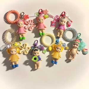 May include: A collection of colorful keychains featuring small doll figures in various outfits. The keychains include pink, yellow, and blue dolls, with accessories like hair ties and charms. The background is white.
