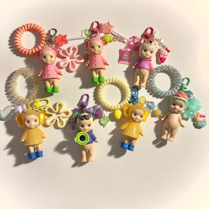 May include: A collection of colorful keychains featuring small doll figures in various outfits. The keychains include pink, yellow, and blue dolls, with accessories like hair ties and charms. The background is white.