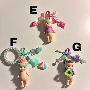 May include: Three small figurines with attached keychains. One figurine wears a pink hoodie and a dog-eared hat, another has a cupcake hat, and the third is a pug with a blue shirt and a green disc.