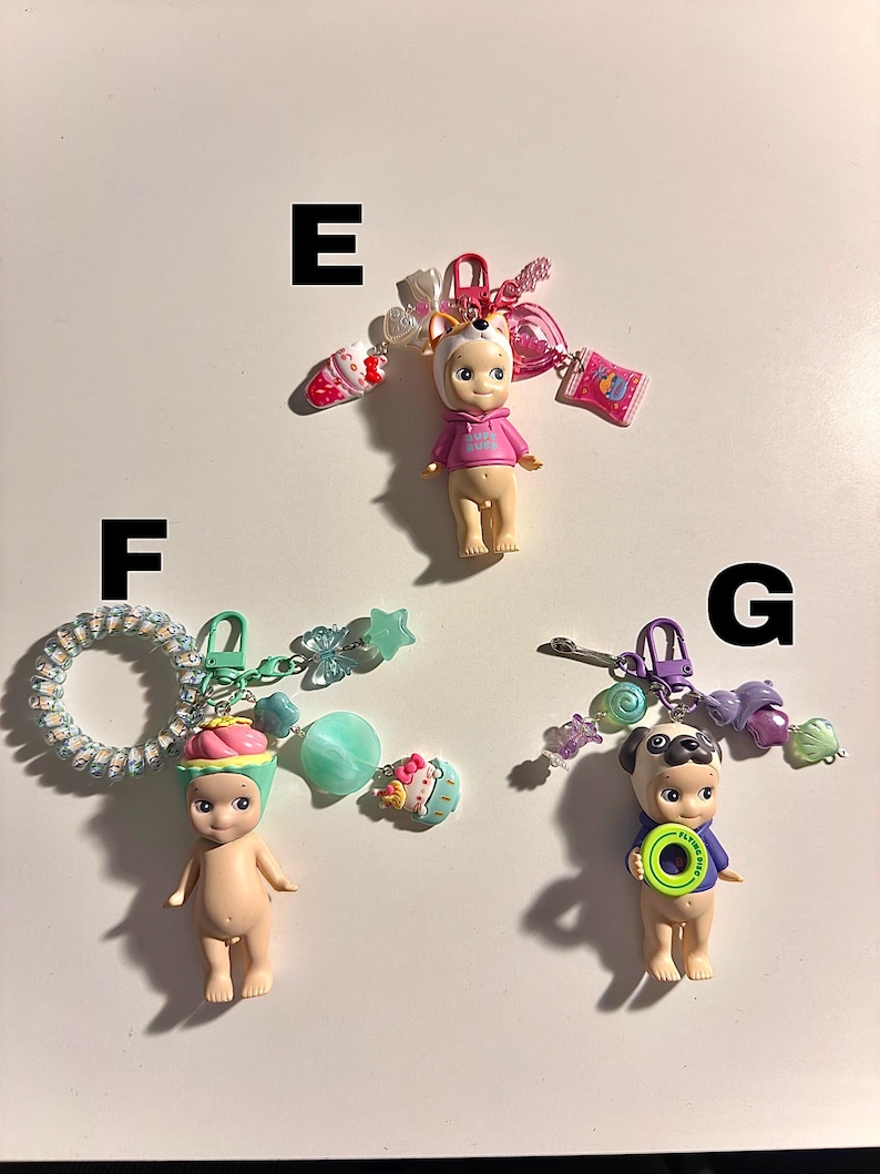 May include: Three small figurines with attached keychains. One figurine wears a pink hoodie and a dog-eared hat, another has a cupcake hat, and the third is a pug with a blue shirt and a green disc.