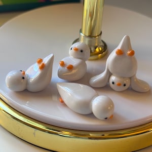May include: Four small, white ceramic duck figurines with orange beaks and feet are arranged on a white and gold-rimmed plate. The ducks are in various poses, some upright and others lying down. The figurines are on a white surface.