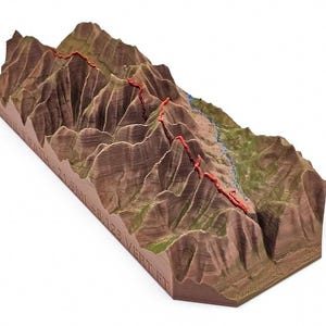 May include: A 3D-printed topographical model of a mountain range, featuring detailed ridges and valleys in shades of brown and green. A red trail winds across the peaks, with a blue river. The base of the model has engraved text.