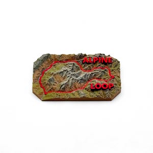 May include: A rectangular 3D topographic relief map of a mountain range featuring a textured brown and green landscape. A bright red line traces a path across the terrain, and the red text "ALPINE LOOP" is prominently displayed on the surface.