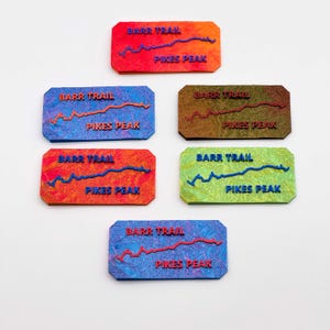 May include: Rectangular plaques with the text "BARR TRAIL" and "PIKES PEAK" in various colors. Each plaque features a raised line design. The plaques are in shades of red, orange, blue, green, and brown.