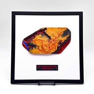 May include: Framed artwork featuring a topographical map of Hell's Revenge 4x4 Trail in Moab, Utah. The map is rendered in shades of orange, red, and blue, set against a white background within a black frame. The text "HELL'S REVENGE 4X4 TRAIL MOAB, UTAH" is displayed below.
