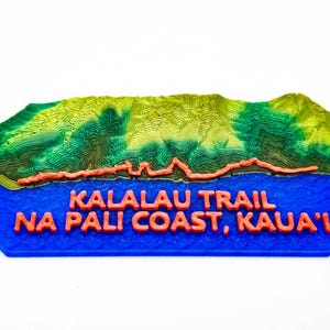 May include: A 3D-printed topographical map of the Kalalau Trail on the Na Pali Coast, Kauai. The map features a blue base with orange lettering and a detailed green and yellow landscape. The text reads "KALALAU TRAIL NA PALI COAST, KAUA'I."