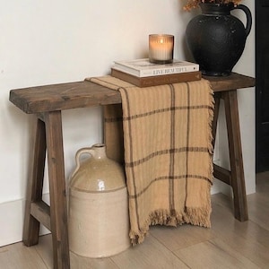 May include: A rustic wooden bench with a plaid throw, books, and a lit candle. A large black vase holds dried flowers. A beige stoneware jug sits below the bench. The scene evokes a cozy, farmhouse aesthetic.