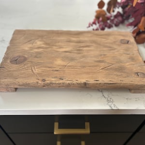 Puede incluir: Una tabla de servir rectangular de madera r&uacute;stica con un acabado natural y envejecido. La tabla presenta vetas de madera visibles, nudos y cabezas de clavos, y se eleva ligeramente de la superficie mediante peque&ntilde;os pies. La tabla mide aproximadamente 46 cm de largo y 30 cm de ancho.