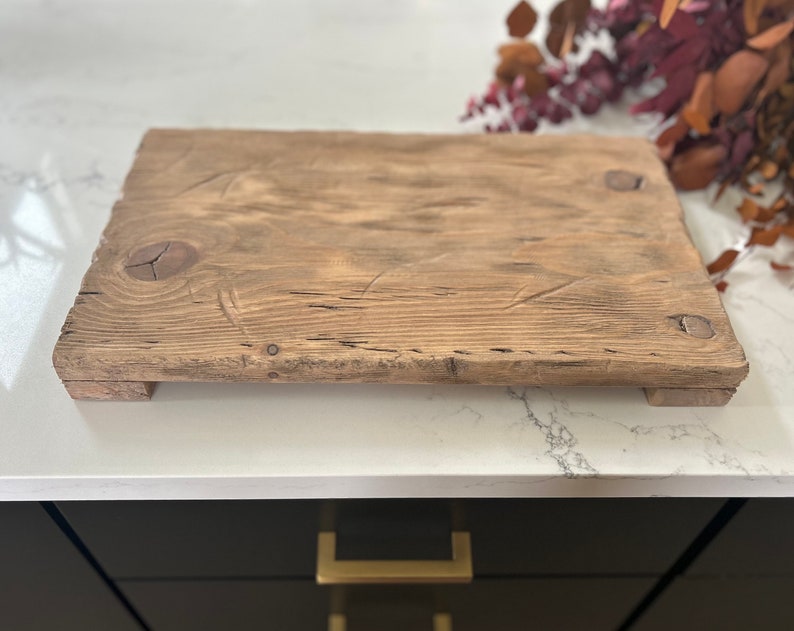 Puede incluir: Una tabla de servir rectangular de madera r&uacute;stica con un acabado natural y envejecido. La tabla presenta vetas de madera visibles, nudos y cabezas de clavos, y se eleva ligeramente de la superficie mediante peque&ntilde;os pies. La tabla mide aproximadamente 46 cm de largo y 30 cm de ancho.