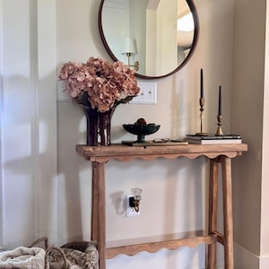 Peut inclure: Une console en bois avec un tablier festonné et quatre pieds droits. Sur la table se trouvent un vase marron foncé avec des hortensias roses séchés, un bol noir, deux bougies noires dans des bougeoirs en laiton et des livres sous un miroir rond.