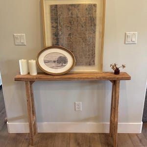 Op de afbeelding: Een rustieke houten console met een ingelijste landschapsafdruk en een grote ingelijste abstracte afdruk. Twee witte pilaar kaarsen en een kleine vaas met gedroogde bloemen staan op de tafel. De tafel is gemaakt van lichtgekleurd hout.