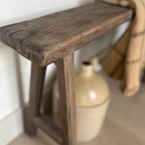 May include: A rustic wooden bench with a weathered, brown finish. The bench has a rectangular seat and angled legs, suggesting a farmhouse or vintage style. A tan blanket is draped over the bench. A beige jug is in the background.