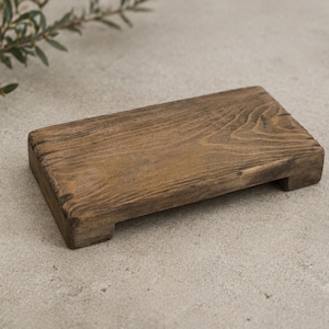 May include: A rectangular, wooden serving board with a natural, distressed finish. The board is a warm brown color and has a slightly raised edge. It rests on two small feet, and is ideal for serving appetizers or small dishes.