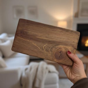 May include: A rectangular wooden cutting board with a natural wood grain pattern. The board is a warm brown color and features a prominent knot detail. The board is held in a person's hand.