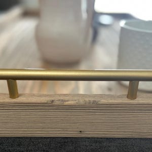 以下が含まれることがあります： 明るい木製の表面に取り付けられた、2つの垂直ポストを備えた、金色の円筒形のキャビネットプルハンドル。 ハンドルはフレームの中央にあります。 白いマグカップとピッチャーが背景にあります。