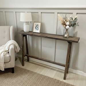 Puede incluir: Una consola rústica de color marrón oscuro con una mesa larga y estrecha y patas anguladas. Una lámpara blanca, obras de arte enmarcadas y un jarrón con flores secas y vegetación se exhiben sobre la mesa. La mesa mide aproximadamente 152 cm de largo.