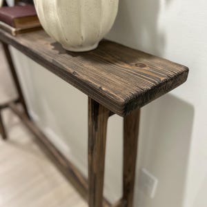 Op de afbeelding: Een rustieke, donker gebeitste houten consoletafel met een lange, smalle bovenkant en vier stevige poten. Een crèmekleurige, getextureerde vaas staat op de tafel. De tafel is gemaakt van massief hout en heeft een natuurlijke, verouderde uitstraling.