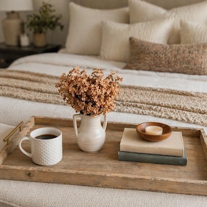 May include: A wooden serving tray with gold handles holds a white mug of coffee, a small vase of dried flowers, a stack of books, and a wooden bowl with a lit candle. The tray sits on a neutral-toned upholstered surface, with a bed in the background.