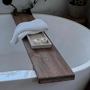 May include: A dark brown wooden bathtub tray with a white waffle-weave towel, a small rectangular tray, and two round objects. The tray spans the width of a white bathtub, creating a spa-like atmosphere.