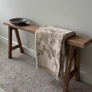 Peut inclure: Banc en bois rustique avec une finition naturelle, comprenant un bol gris foncé et une couverture beige avec un motif floral. Le banc est soutenu par des pieds en forme de A, mesurant environ 150 cm de long.