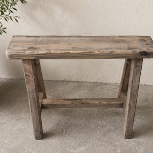 May include: A rustic wooden bench with a rectangular seat and angled legs. The bench is made of weathered wood, showcasing natural grain and knots. The bench is approximately 76 cm long.
