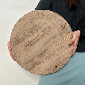 May include: A round, unfinished wooden tabletop with a natural, weathered appearance. The wood grain is visible, and the surface has a rustic, aged look. The tabletop is held by a person.