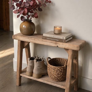 May include: A rustic wooden bench with a lower shelf, holding a pair of tan boots and a woven basket. The top shelf displays a brown vase with burgundy foliage, books, and a lit candle. The bench is made of light-colored wood.