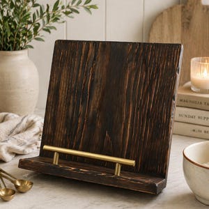 Puede incluir: Un atril de madera marr&oacute;n oscuro para libros de cocina con una barra met&aacute;lica dorada. El atril tiene un aspecto r&uacute;stico y texturizado y est&aacute; dise&ntilde;ado para sostener libros de cocina o tabletas. Un accesorio de cocina decorativo.
